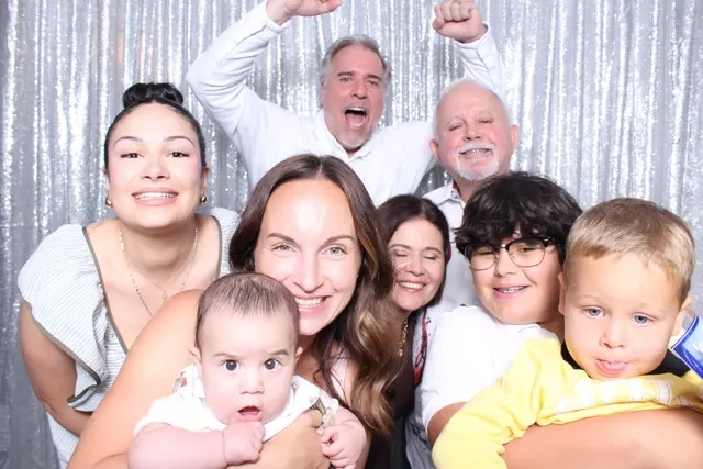 Multigenerational family group, including two young children, in front of a silver sequin backdrop.