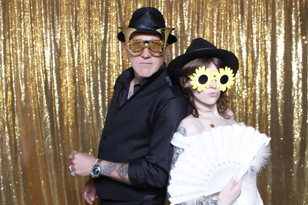 Two wedding guests posing back-to-back with hats, novelty glasses, and a feather fan
